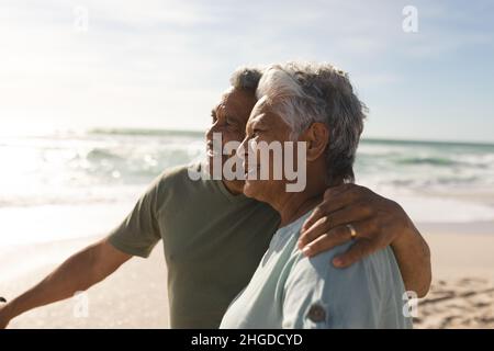 Älterer Biracial-Mann mit Arm um die Frau, die am Fahrrad steht und an sonnigen Tagen auf den Strand blickt Stockfoto