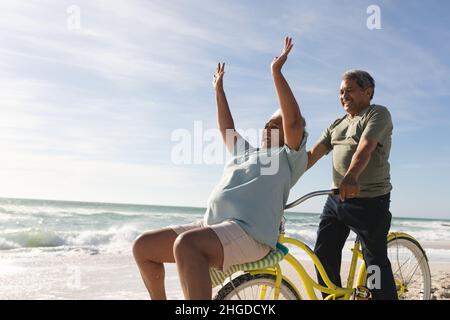 Fröhliche ältere Frau aus dem Birazial jubelt, während der Mann am sonnigen Tag am Strand Fahrrad fährt Stockfoto