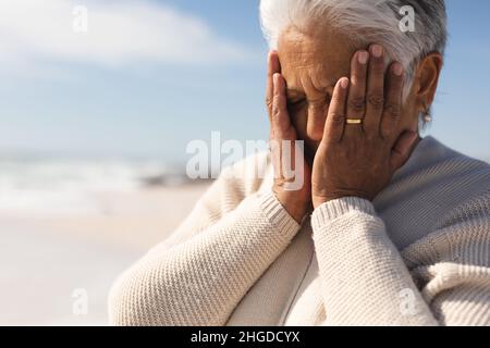 Besorgte pensionierte ältere Birazialfrau mit Händen, die das Gesicht am Strand an einem sonnigen Tag bedeckt Stockfoto