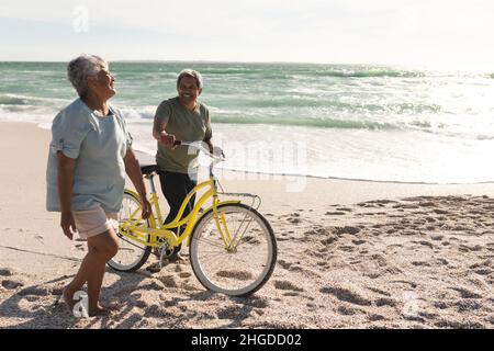 Fröhliche ältere Frau aus dem Birazial, die an sonnigen Tagen mit einem Mann am Strand mit dem Fahrrad unterwegs ist Stockfoto