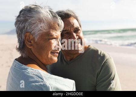 Nachdenkliches mehrrassiges Senioren-Paar, das an einem sonnigen Tag am Strand steht und zusammen wegschaut Stockfoto