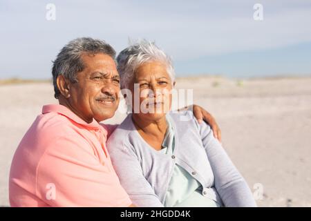 Nachdenkliches, multirassisches Seniorenpaar, das sich an einem sonnigen Tag am Strand entspannt Stockfoto