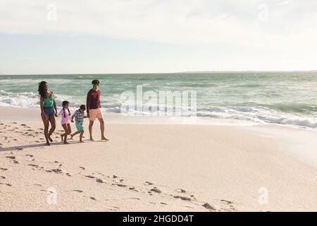 Die ganze Länge der multirassischen Familie hält die Hände, die am sonnigen Strand mit Fußabdrücken an der Küste spazieren gehen Stockfoto