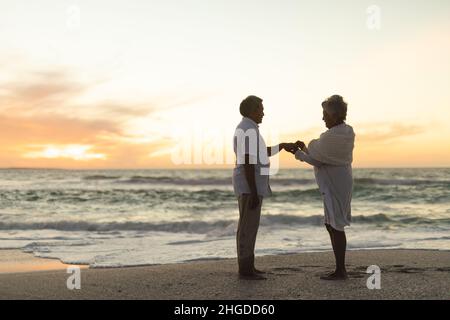 Seitenansicht der älteren biracial Braut, die Ehering auf den Finger des Bräutigams am Strand während des Sonnenuntergangs setzt Stockfoto