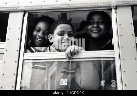 Eine fröhliche Familie blickt aus einem Schulbusfenster, während sie ein Kaninchenohrschild aufblitzen. In Brooklyn, New York um 1976. Stockfoto