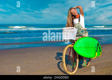 Junge Frau mit Surfbrett und Fahrrad am Strand. Stockfoto