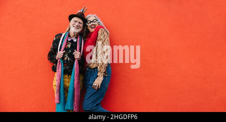 Ein älteres Paar lacht fröhlich, während es vor einem roten Hintergrund zusammensteht. Fröhliches Seniorenpaar, das sich in farbenfroher Freizeitkleidung lebhaft fühlt. Stockfoto