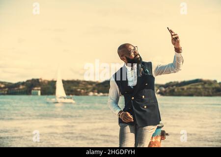 Ein eleganter afroamerikanischer Senior in modischem Outfit sitzt auf einem Poller neben dem Fluss und hat an einem schillernden Tag einen Videoanruf; ein eleganter Stockfoto