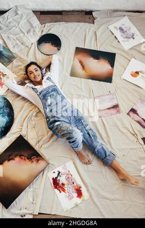 Draufsicht auf eine Künstlerin, die sich in der Mitte ihrer Bilder hinlegt. Fröhliche junge Malerin lächelt in ihrer Kunstgalerie vor der Kamera. Kreative junge wo Stockfoto