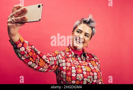 Nehmen Sie ein niedliches Selfie. Glückliche junge Frau lächelt fröhlich, während sie für ein Selbstporträt auf ihrem Smartphone posiert. Junge Frau mit gefärbtem Haar stehend aga Stockfoto