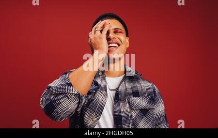 Stilvoller junger Mann, der fröhlich lächelt und vor einem roten Hintergrund steht. Modischer junger Mann mit Nagellack und Lidschatten in einem Studio. Nein Stockfoto
