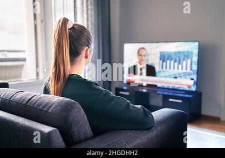 TV-Nachrichten. Frau, die im Wohnzimmer Fernsehen schaut. Reporter, Anchor und Studio-Moderator auf dem Bildschirm mit Nachrichtenüberschriften. Sitzende Person. Stockfoto