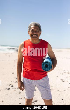 Porträt eines lächelnden pensionierten Birazialmannes, der am Strand eine gerollte Yogamatte gegen den blauen Himmel hält Stockfoto