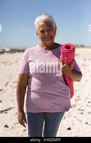 Porträt einer lächelnden älteren Birazialfrau im Ruhestand, die am Strand eine Yogamatte mit kurzen weißen Haaren hält Stockfoto