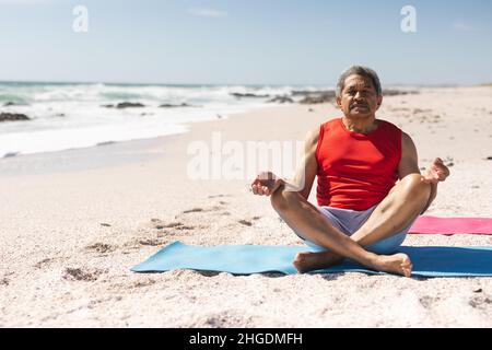 Die ganze Länge eines älteren Birazialmannes in Lotusposition, der auf einer Yogamatte am sonnigen Strand meditiert Stockfoto