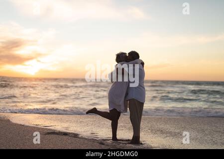 Seitenansicht eines mehrrassigen älteren Ehepaares, das sich während des Sonnenuntergangs am Strand umarmte und küsste Stockfoto