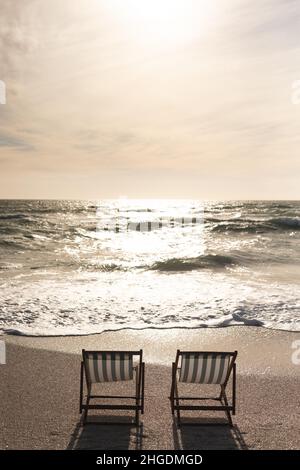 Zwei leere Klappstühle aus Holz am Ufer vor Wellen, die am Strand gegen den Himmel im Meer plätschern Stockfoto