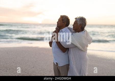 Seitenansicht eines mehrrassigen älteren Ehepaares, das während des Sonnenuntergangs am Strand umarmt und wegschaut Stockfoto