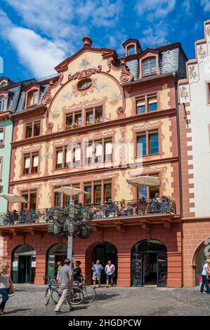 Wunderschöne Aussicht auf ein Gebäude auf dem Marktplatz in der Altstadt von Mainz. Die Fassade hat eine schöne einheimische Wandmalerei und auf dem Balkon Menschen... Stockfoto