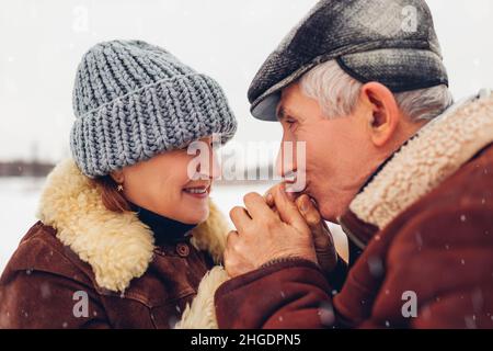 Ein glückliches Ehepaar aus der älteren Familie spaziert im verschneiten Winterwetter im Park. Älterer, fürsorglicher Mann wärmt die Hände der Frau mit Atem. Valentinstag Stockfoto