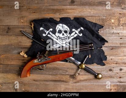 Gekreuzte antike Feuerstein-Pistole und großer Piratendolch und fröhliche roger-Flagge auf einem rauen Holztisch Stockfoto