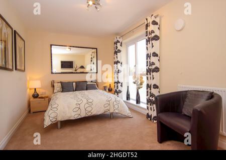 Schlafzimmer in neutralen Farbtönen, Doppelbett mit Kingsize-Bett, niedrige Fensterbank, Vorhänge mit Blattmuster, Spiegelwand, beigefarbener Teppich. Stockfoto