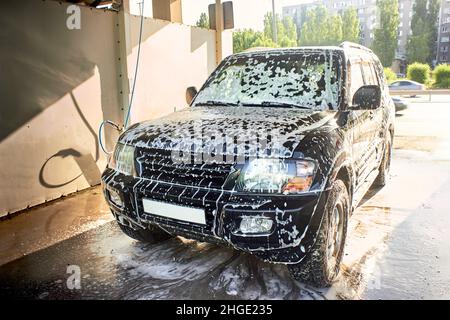 Ein schwarzer SUV-Wagen bei einer Autowaschanlage zur Selbstbedienung. Waschen Sie das Auto mit Ihren eigenen Händen. Ein schwarzes Auto in Reinigungsschaum. Stockfoto