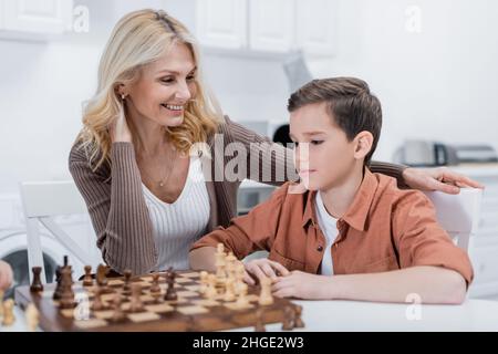 Frau mittleren Alters lächelt in der Nähe des Enkels, der in der Küche Schach spielt Stockfoto