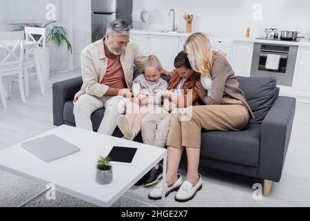 Paar haben Spaß mit Enkeln, während sie auf der Couch in der Nähe von Laptop und digitalem Tablet auf dem Tisch sitzen Stockfoto