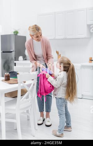 Lächelnde Frau hält die Lunchbox, während sie den Rucksack der Enkelin in der Küche packt Stockfoto