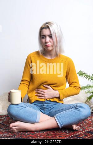 Junge Frau, die zu Hause sitzt und eine Tasse Tee hält, während sie ihre Hand in ihren Magen legt Stockfoto