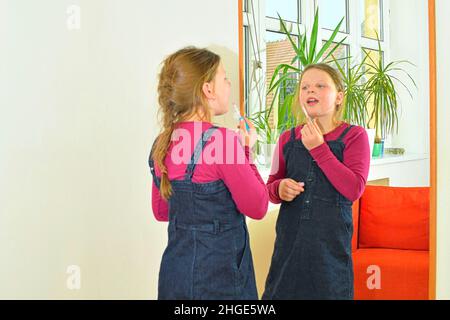 Beauty- und Modekonzept. Kind mit schönen Wangen lächelt und malt drinnen Lippen. Kleines Mädchen hält Lippenstift oder Mascara vor dem Spiegel in der Stockfoto