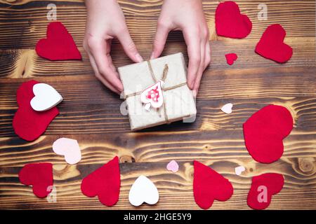 Hände, die eine Schachtel aus Kraftpapier mit herzförmigem Dekor auf einem Hintergrund halten, der zum Valentinstag dekoriert ist. Geschenk für ein geliebtes Konzept. Weichfokus Stockfoto