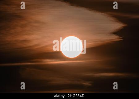 Leicht bewölkt, rot getönter Himmel. In der Mitte sieht man eine helle Sonnenscheibe. Stockfoto
