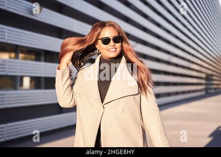 Afroamerikanische Frau mit Sonnenbrille. Die erfolgreiche junge Frau trägt einen Geschäftsanzug. Sie geht kraftvoll, während sie ihre Haare zu einem kämmt Stockfoto