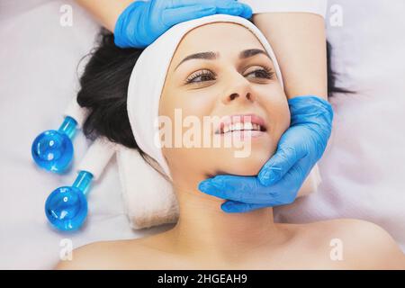 Kosmetikerin in blauen Handschuhen, die den Kopf der Frau mit zwei Händen und einem Massageinstrument für die Gesichtswalze in der Nähe des Tisches hält. Eisballse Stockfoto