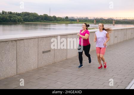 Breite Aufnahme von persönlichen Fitness weibliche Trainer hilft fetten Frau Gewicht zu verlieren, außerhalb Laufen entlang der Stadt am Wasser im Sommer Morgen. Stockfoto