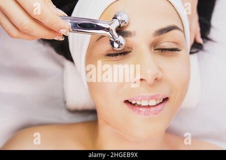 Oben Foto einer schönen fröhlichen glücklichen Frau, die in einem Schönheitssalon eine Gesichtsmassage mit Rollerausrüstung von einem Kosmetiker erhält. Kaukasische Brünette Stockfoto