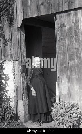 1950s, historische, ältere Bauernhofdame, die eine lange Schürze über einem knöchellangen Rock trägt und in Pantoffeln am Eingang einer Holzscheune steht, Prynees, Frankreich. Stockfoto