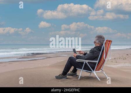 Ein erwachsener Mann sitzt auf einem Strandstuhl auf einem medano am Strand und tippt auf dem Smartphone. Horizontales Foto Stockfoto