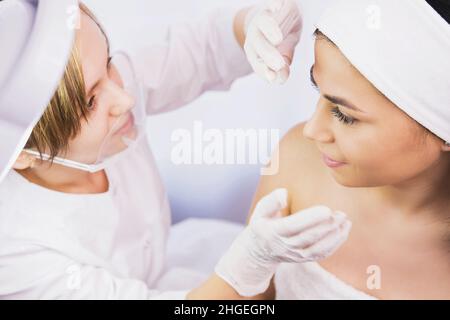 Draufsicht Foto einer Klientin, die in der Schönheitsklinik einer medizinischen Untersuchung des Gesichts durch eine Kosmetikerin mit Handschuhen unterzogen wird. Stockfoto