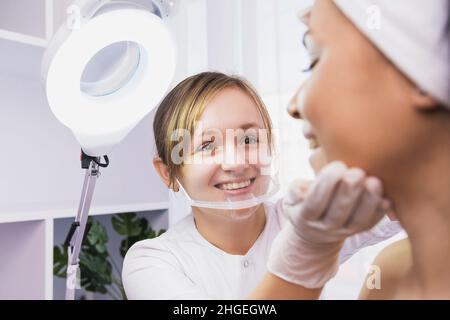 Professionelle Kosmetologin Frau trägt Gesichtsschutzmaske macht medizinische Klinik Gesicht Untersuchung einer Frau nach der Verjüngung Verfahren in Stockfoto