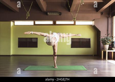 Die blonde Frau, die Yoga praktiziert, führt eine Variation der Ardha chandrasana-Übung, Halbmondpose durch und steht im Studio auf einer Matte Stockfoto