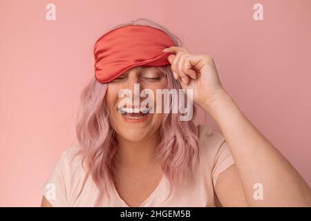 Positive Frau lächelt zahnhaft zieht rote Seide Schlafmaske erwacht in guter Stimmung, nachdem sie angenehme Träume in der Nacht genießt Morgenzeit gegen zu sehen Stockfoto