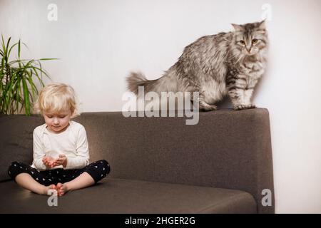 Nettes kleines Mädchen sitzt mit Beinen gefaltet auf der Couch im Haus. Gesunder Lebensstil. Kleinkindsport. Das Kind macht am Morgen danach Entspannungsübungen Stockfoto