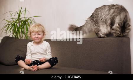 Nettes kleines Mädchen sitzt mit Beinen gefaltet auf der Couch im Haus. Gesunder Lebensstil. Kleinkindsport. Das Kind macht am Morgen danach Entspannungsübungen Stockfoto