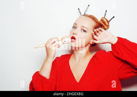 Schönes Mädchen, das Sushi auf einem weißen Teller isst. Eine Frau in einem roten Kleid und roten Lippen isst Sushi-Essstäbchen mit Lachs und Garnelen. Mode Fotomodell Stockfoto