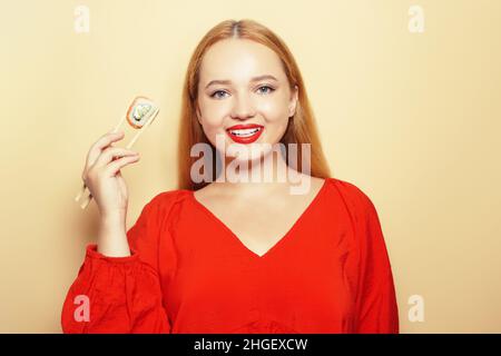 Schönes Mädchen, das Sushi auf einem weißen Teller isst. Eine Frau in einem roten Kleid und roten Lippen isst Sushi-Stöcke mit Philadelphia-Lachs. Model lächelt und Toilette Stockfoto