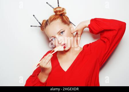 Schönes Mädchen, das Sushi auf einem weißen Teller isst. Eine Frau in einem roten Kleid und roten Lippen isst Sushi-Essstäbchen mit Lachs und Garnelen. Mode Fotomodell Stockfoto