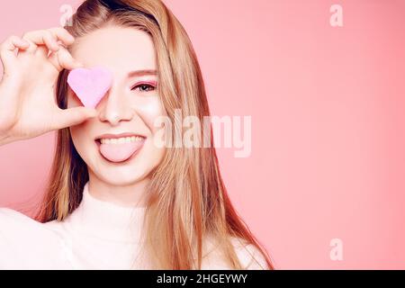 Valentine's Day, ein Mädchen auf einem rosa Hintergrund, schließt ihr Auge mit einem Herzen. Die Blondine lächelt schön, lacht, zeigt ihre Zunge, macht Gesichter, hat f Stockfoto